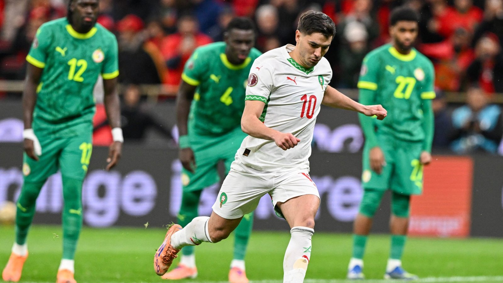Moroccan player wearing jersey number 10 takes a penalty kick, with Senegalese defenders in green kit looking on in the background.