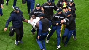 Security personnel carry a player off the pitch amid a chaotic scene, wearing navy jackets and caps, during a tense football match.