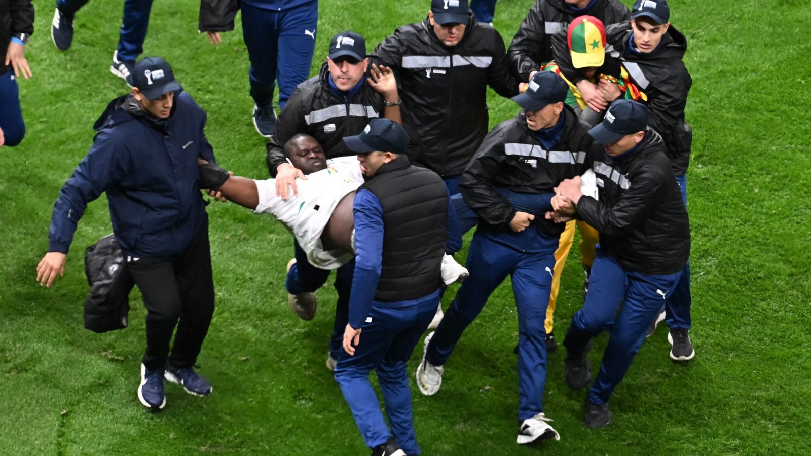 Security personnel carry a player off the pitch amid a chaotic scene, wearing navy jackets and caps, during a tense football match.