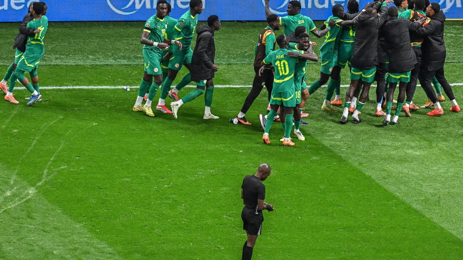 Senegal players in green kits celebrate a goal with excitement on the pitch, while the referee stands nearby observing the scene.