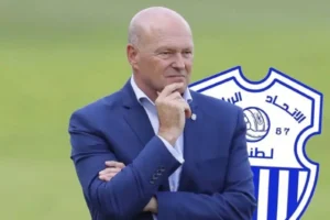 Former coach in a blue suit contemplating, with the logo of Ittihad Tanger in the background, representing Moroccan football.
