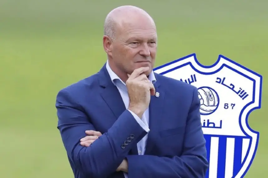 Former coach in a blue suit contemplating, with the logo of Ittihad Tanger in the background, representing Moroccan football.