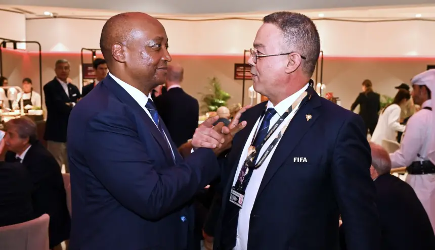 Twee functionarissen voeren een gesprek tijdens een formeel evenement, gekleed in marineblauwe pakken en voorzien van FIFA-accreditatie, wat de bestuurlijke kant van het voetbal benadrukt.