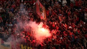 Al-Ahly fans in red jerseys celebrate passionately, holding a flag and creating a dramatic scene with smoke during a match.