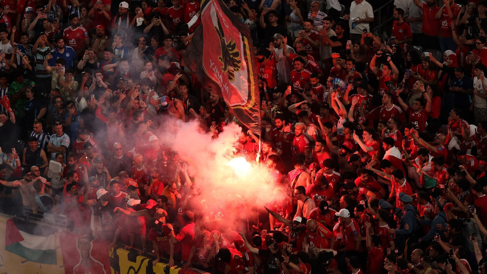 Al-Ahly fans in red jerseys celebrate passionately, holding a flag and creating a dramatic scene with smoke during a match.