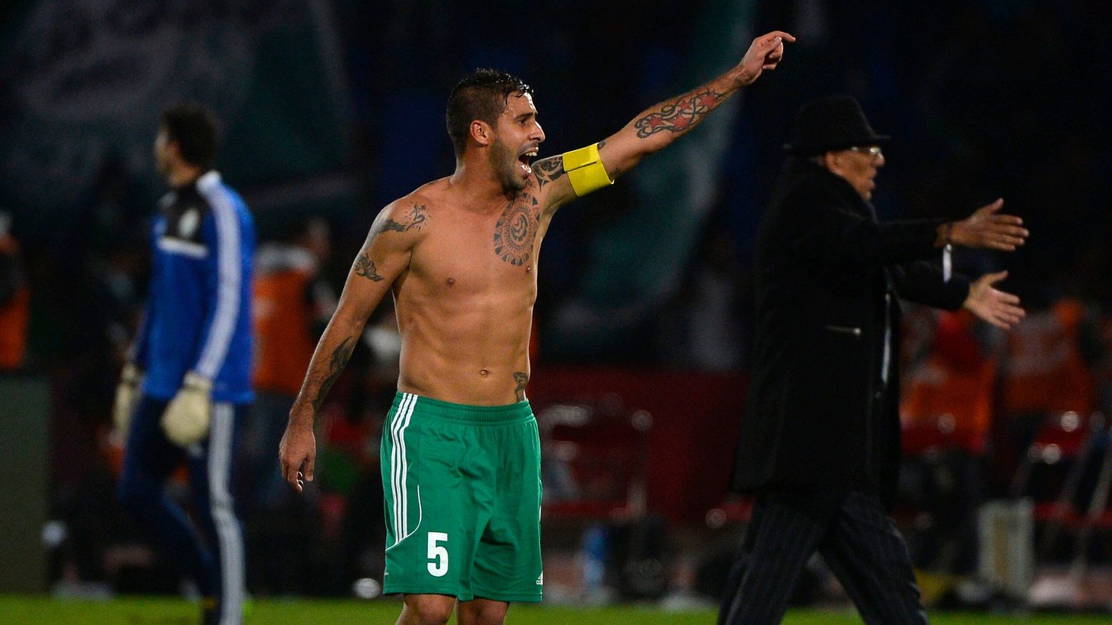 Mohsen Moutouali, wearing green shorts and a captain's armband, celebrates passionately shirtless during a Raja Casablanca match.