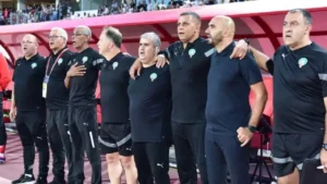 Moroccan football coaching staff stand together in black outfits, singing the national anthem during a match, showcasing team unity.