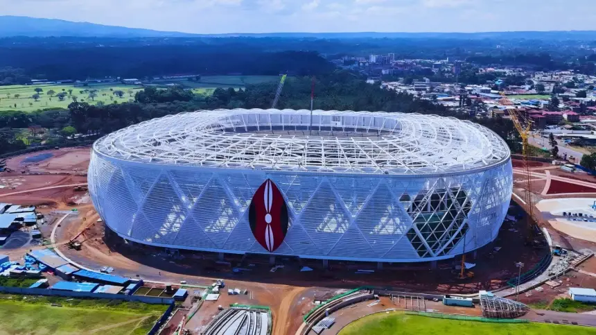 Vue aérienne d'un stade moderne au design distinctif, présentant une structure en treillis blanc et un emblème rouge, se détachant sur un ciel bleu dégagé.
