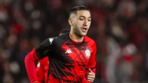 Hakim Ziyech dans un maillot rouge et noir saisissant, démontrant un mouvement dynamique pendant un match, représentant le football marocain avec fierté.