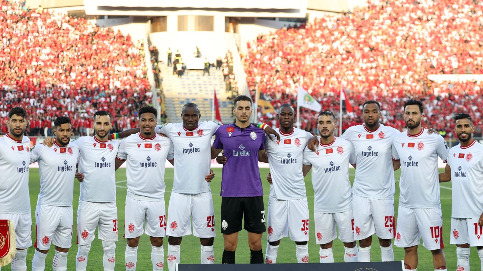 Wydad Casablanca-spelers in witte tenues poseren gezamenlijk voor een teamfoto voorafgaand aan een wedstrijd, met een achtergrond van in het rood geklede fans.