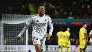Kylian Mbappé celebrates after scoring for Real Madrid, featuring yellow stadium colours and a cheering crowd in the background.