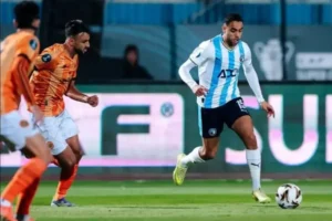 Moroccan players in action during a match; one in a blue and white kit dribbles past an opponent in an orange kit.