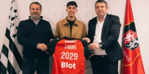 Zabiri holds his new red football jersey with "ZABIRI 2029," flanked by two officials, celebrating his transfer to Rennes.