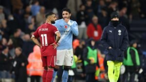 Mohamed Salah in a red Liverpool kit talks to a Manchester City player in blue, set against a stadium backdrop during a match.