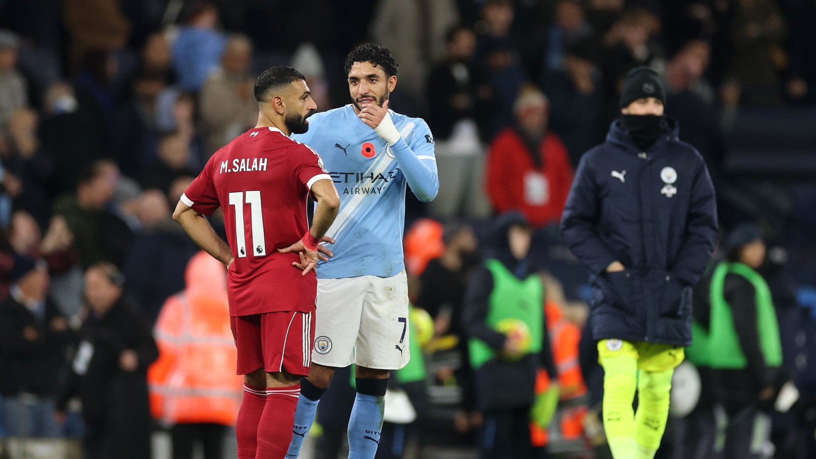 Mohamed Salah in a red Liverpool kit talks to a Manchester City player in blue, set against a stadium backdrop during a match.