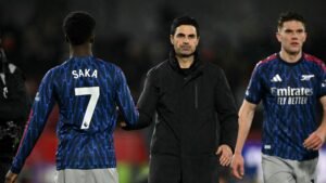 Arsenal's Bukayo Saka in a blue kit with '7' on the back, shaking hands with manager Mikel Arteta, alongside teammate in red Emirates kit.