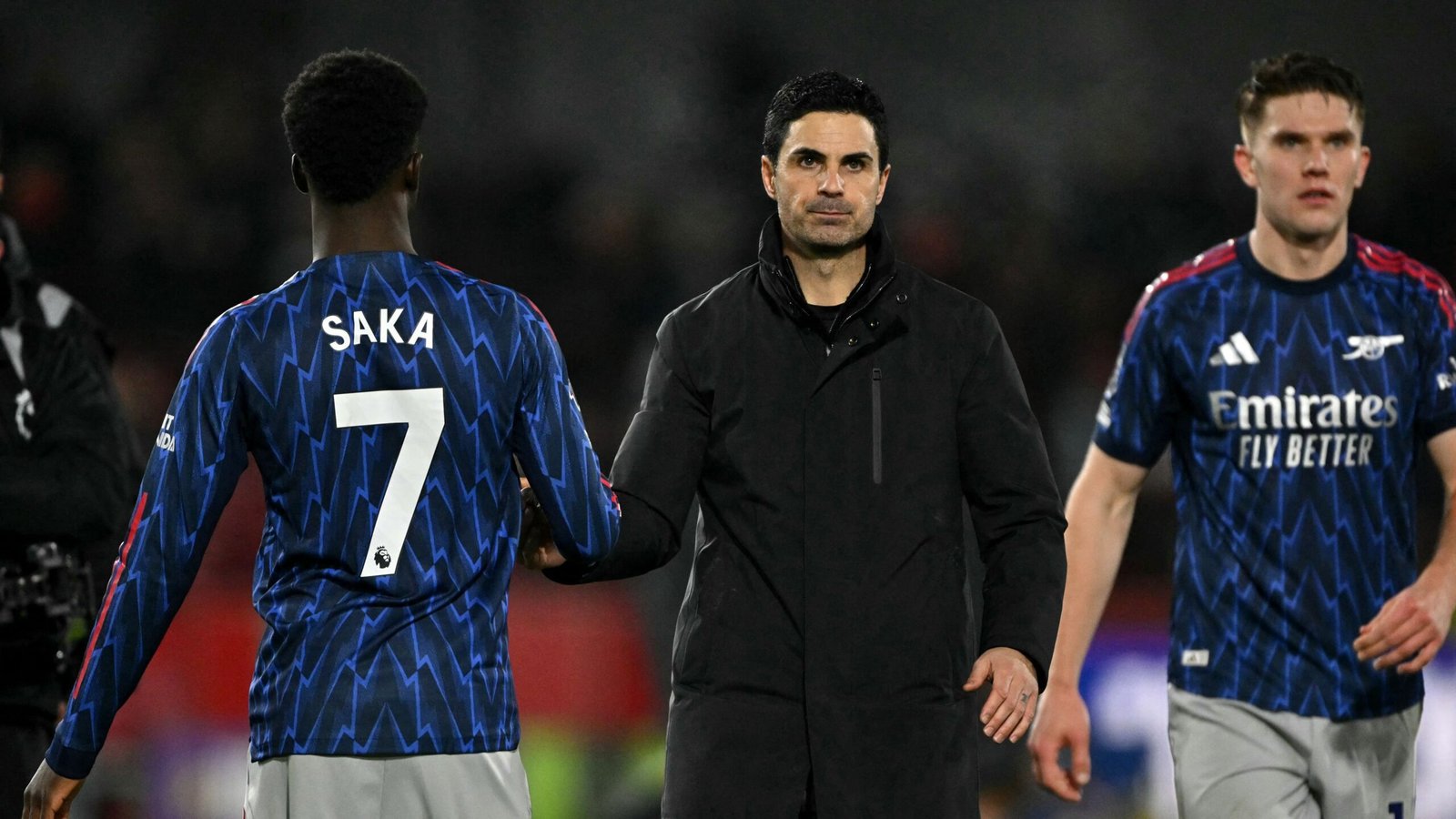 Arsenal's Bukayo Saka in a blue kit with '7' on the back, shaking hands with manager Mikel Arteta, alongside teammate in red Emirates kit.