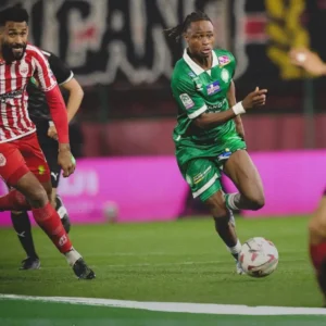 Young Moroccan player in green kit dribbles past an opponent in red during an intense league match, showcasing agility and skill.