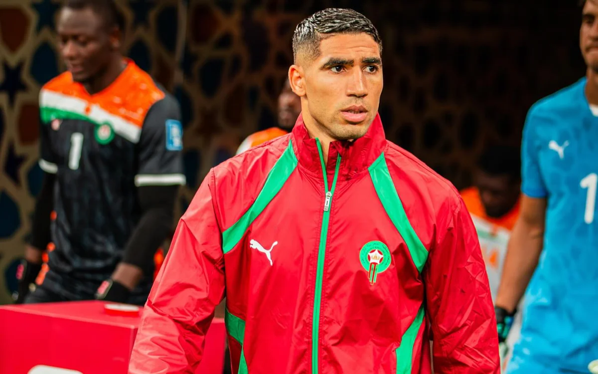 Achraf Hakimi in a red Moroccan tracksuit, focused as he walks out onto the pitch, with goalkeepers in the background during a match.