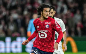 Ayyoub Bouaddi en maillot rouge du Lille OSC, concentré sur le ballon pendant un match, avec un défenseur visible en arrière-plan.