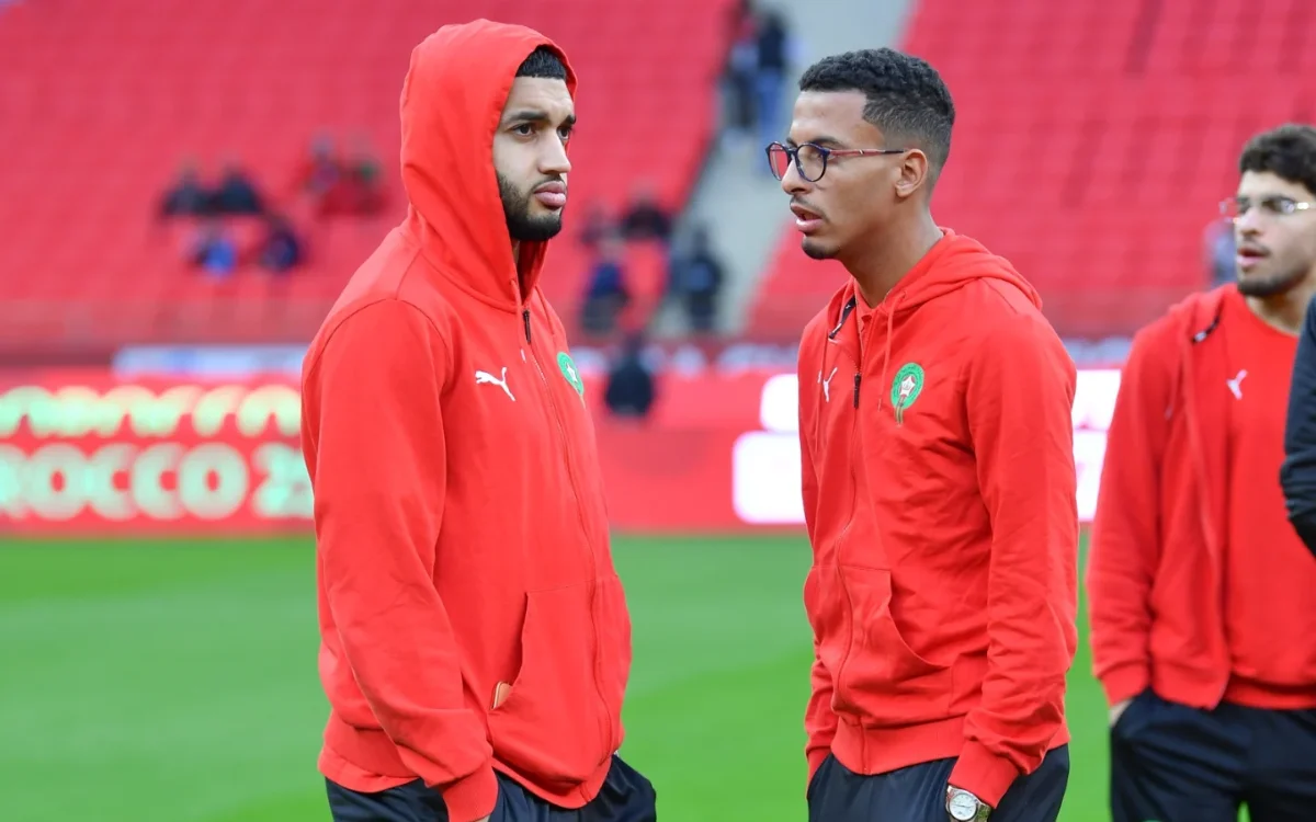 Azzedine Ounahi in gesprek met een teamgenoot, beiden gekleed in rode hoodies, wat hun concentratie toont voor een wedstrijd van het Marokkaans nationaal elftal.