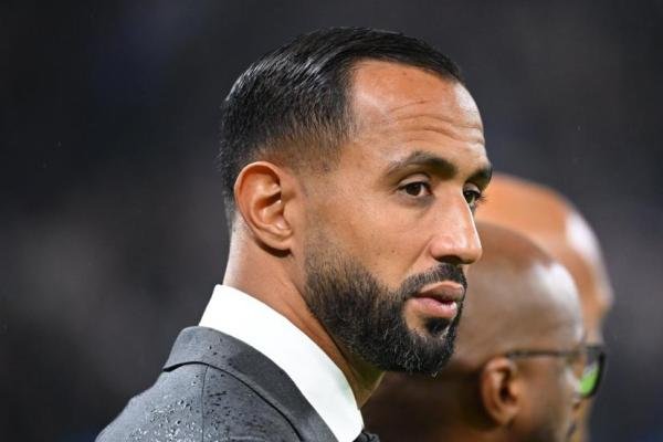 Medhi Benatia in een grijs pak, peinzend opzij kijkend, met een wazig stadion op de achtergrond, wat een moment van bezinning vastlegt.