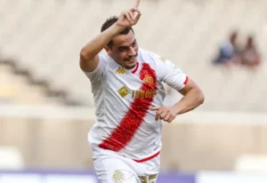Celebrating player Abderrazak Hamdallah in Wydad Casablanca's white and red kit, joyfully pointing skyward after scoring a goal.