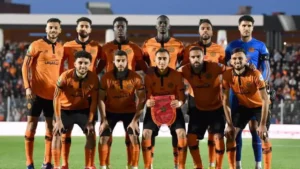 Aoudad-spelers poseren trots in hun oranje-zwarte tenue en tonen het clubembleem voor een levendige stadionachtergrond.