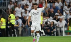 Brahim Diaz in het witte tenue van Real Madrid, terwijl hij zijn hand opsteekt tijdens een wedstrijd in het Santiago Bernabéu Stadion, omringd door fans.