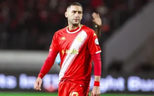 Hakim Ziyech in a red Wydad Casablanca kit, looking focused during a CAF Cup match, with fans blurred in the background.