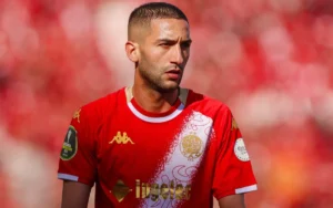 Hakim Ziyech wearing Wydad Casablanca's red kit, focused during a match against a blurred red background of fans.