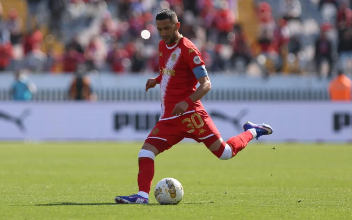 Hakim Ziyech, wearing a red Wydad Casablanca kit with captain's armband, skillfully plays the ball during a match.