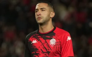 Hakim Ziyech, gekleed in een rood-zwart voetbaltenue, staat geconcentreerd op het veld en toont zijn toewijding aan de sport.