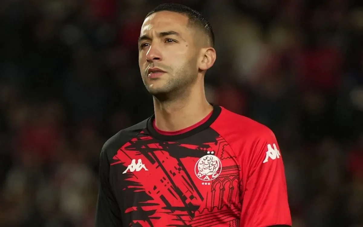 Hakim Ziyech, gekleed in een rood-zwart voetbaltenue, staat geconcentreerd op het veld en toont zijn toewijding aan de sport.