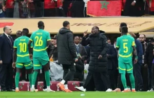 Senegal players, including Sadio Mané, in vibrant green and yellow kits, interact with staff during a match at a stadium, with Moroccan fans visible in the background.