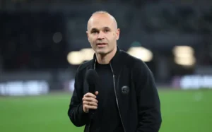 Andrés Iniesta spreekt het publiek toe in een zwart jasje, met een microfoon in de hand, met een vervaagde voetbalveld op de achtergrond.