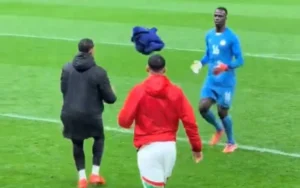 Ismail Saibari, en tenue d'entraînement rouge, lance une veste bleue à un coéquipier, tandis qu'un gardien de but en bleu se prépare sur le terrain.