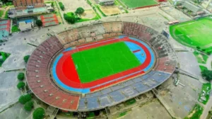 Aerial view of a large football stadium featuring a bright green pitch surrounded by red and blue running tracks, set in a urban environment.