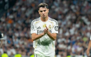 Brahim Diaz in Real Madrid's white kit, clapping after a match, with a blurred crowd in the background, highlighting his focus and dedication.