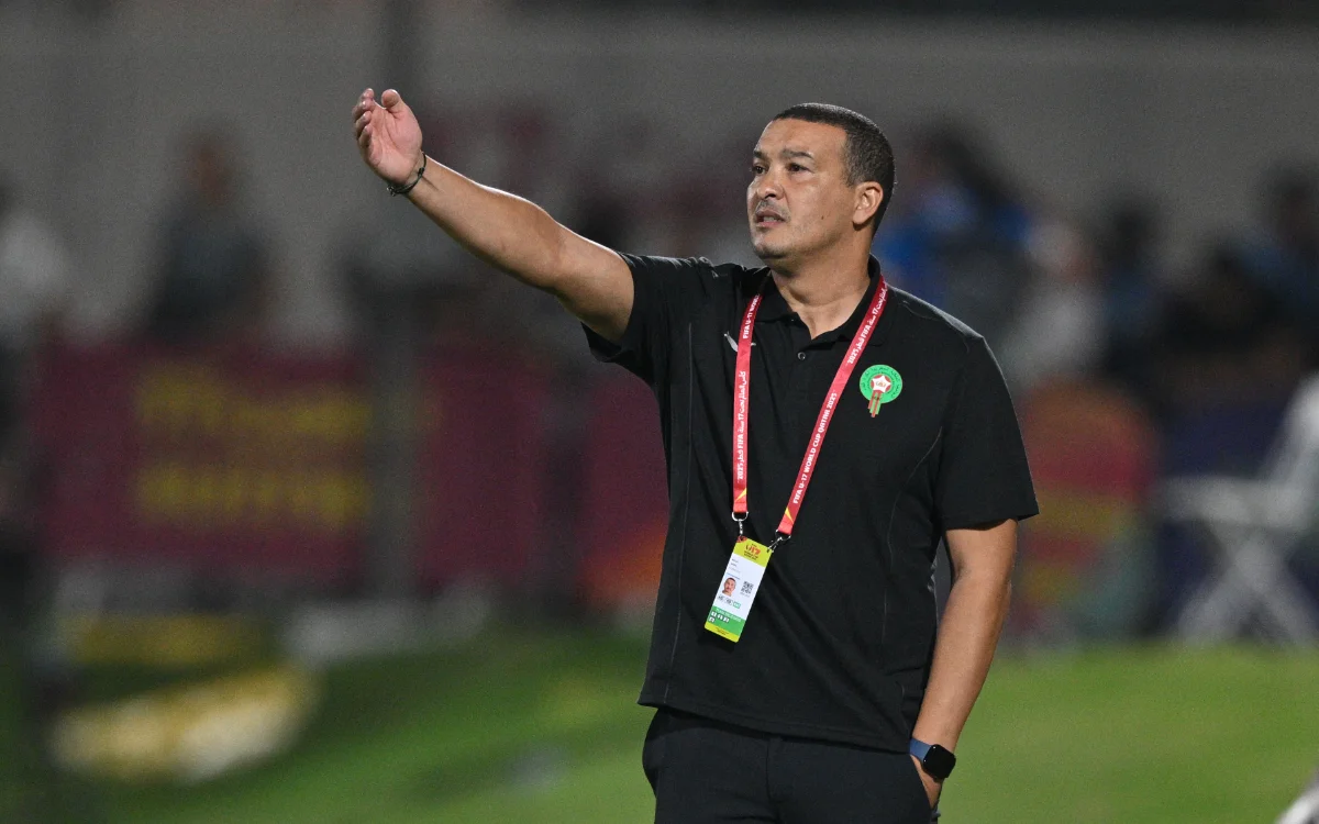 Morocco's football coach, Nabil Baha, giving instructions during a match, wearing a black polo with a team badge and a wristwatch.