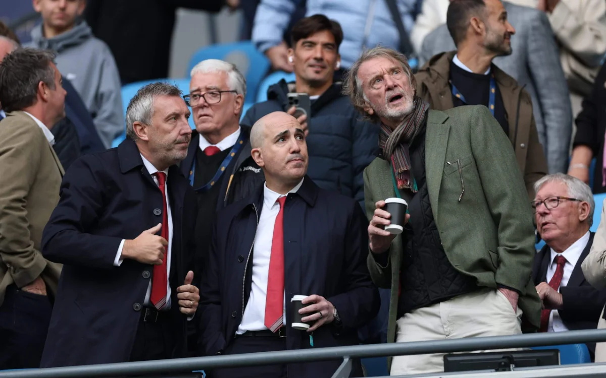 Four men in formal attire, some holding coffee cups, appear engaged and observing a football match from the stands.
