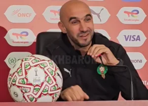 Walid Regragui at a press conference, smiling and holding a football adorned with Moroccan colours, discussing the CAF tournament.