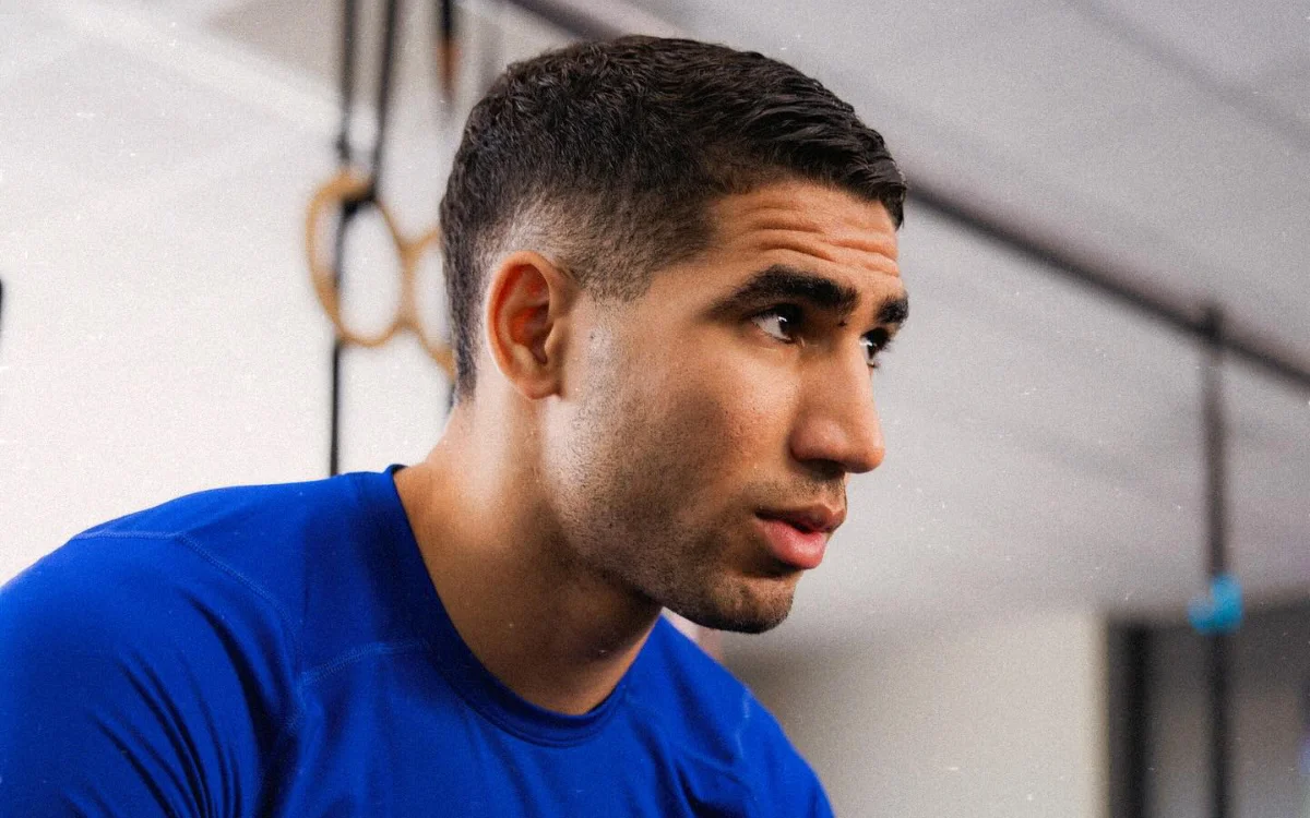 Achraf Hakimi, wearing a blue training shirt, gazes thoughtfully to the side in a gym setting, representing PSG and Moroccan football.
