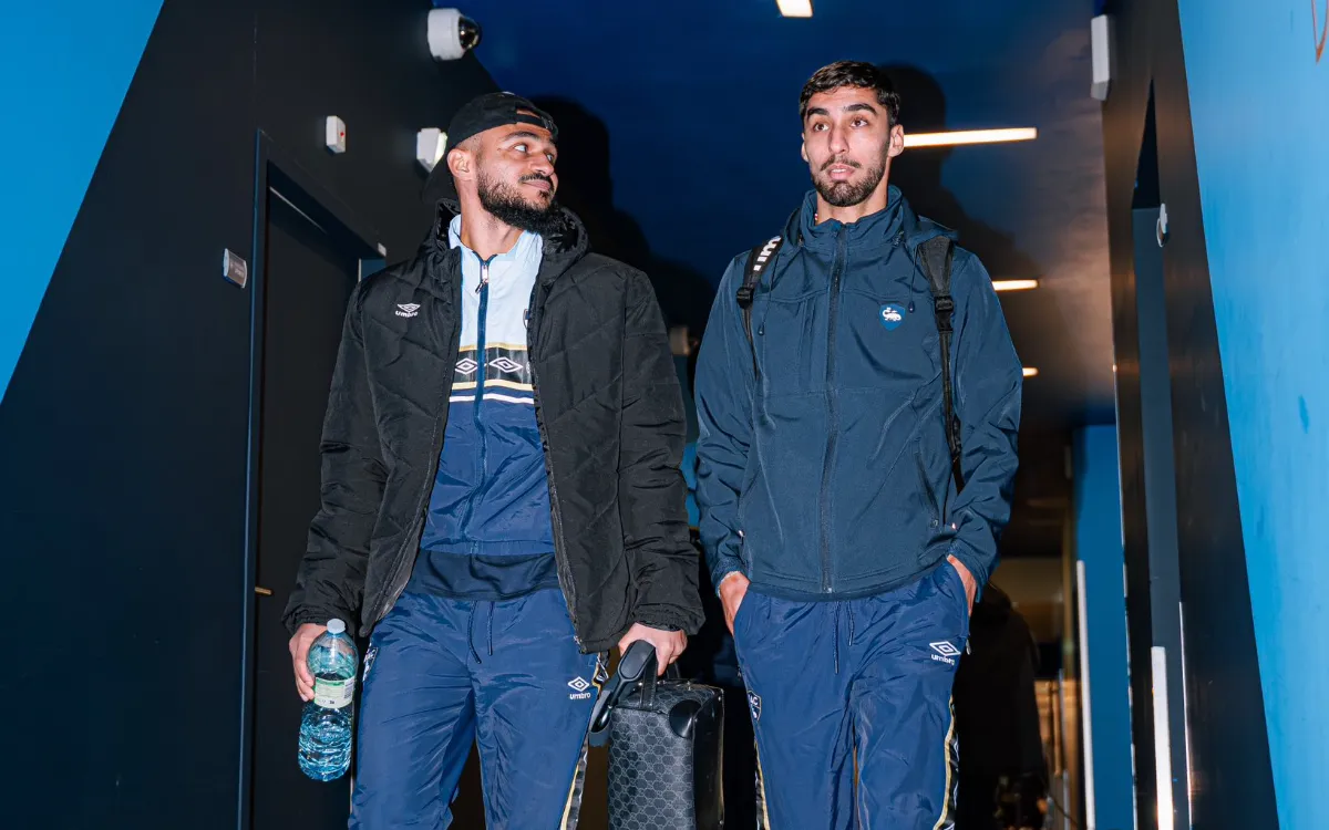 Soufiane Boufal et un coéquipier traversent un hall d'aéroport, vêtus de tenues d'entraînement marine, prêts à voyager.