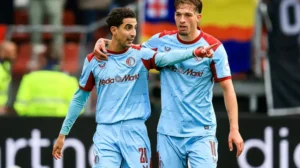 Targhaline and teammate celebrating a goal in their light blue away kit, with fans and team banners in the background.