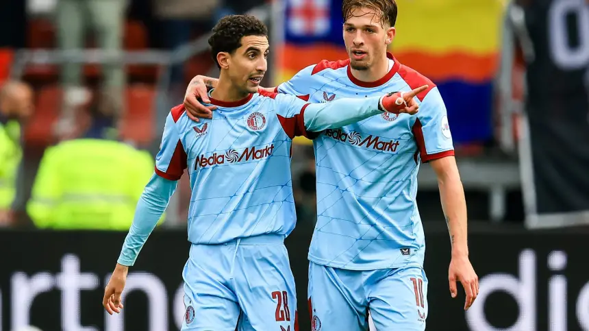 Targhaline and teammate celebrating a goal in their light blue away kit, with fans and team banners in the background.
