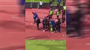 Players and staff from both teams engage in a heated confrontation on the pitch, surrounded by scattered cups and an animated crowd.