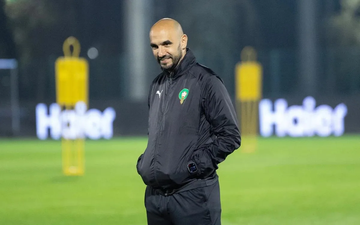 Walid Regragui lachend tijdens een trainingssessie, gekleed in een zwart trainingspak met het embleem van Marokko, omringd door trainingsmateriaal.