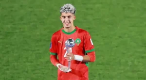 Zabiri, vêtu d'un maillot rouge du Maroc, sourit en tenant un trophée sur le terrain, célébrant une réussite footballistique.